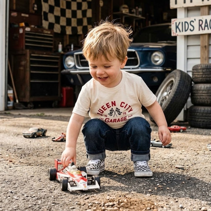 A young boy wearing a cream graphic t-shirt and jeans crouches on a gravel driveway in front of a garage, playing with a small toy race car. The garage background features a blue classic car, toolboxes, and a sign that reads 'PIT ROW KIDS RACING'.