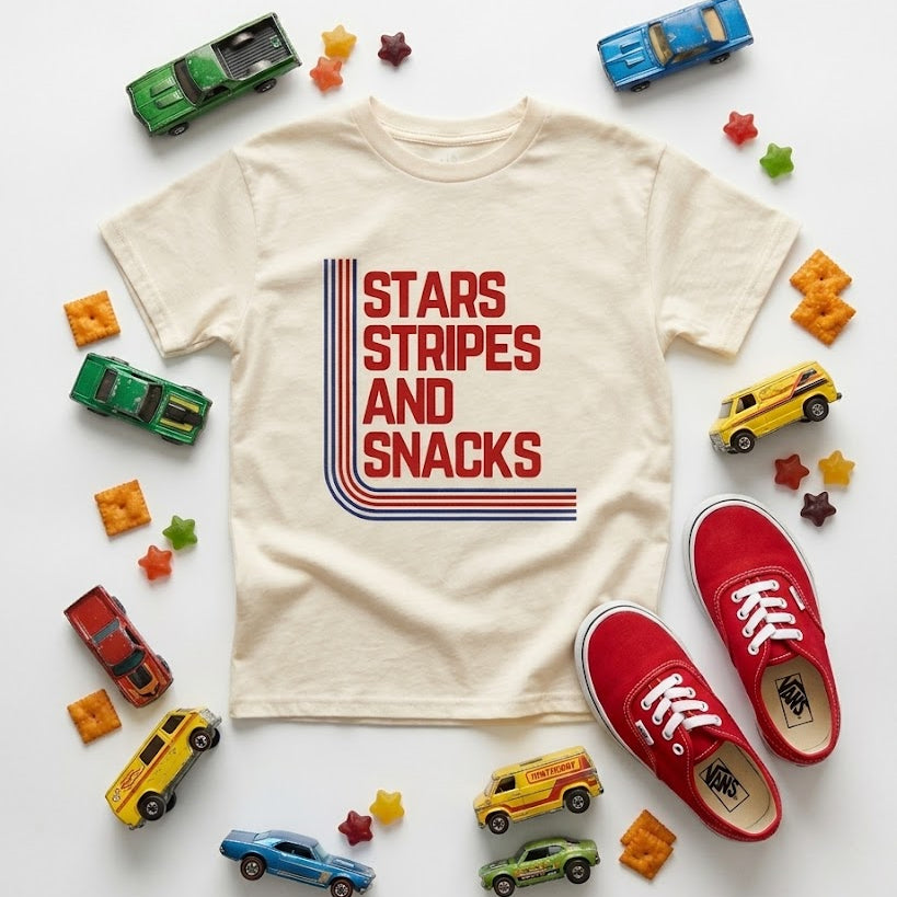 Beige t-shirt with 'Stars Stripes and Snacks' text, red sneakers, and toy cars on a white background