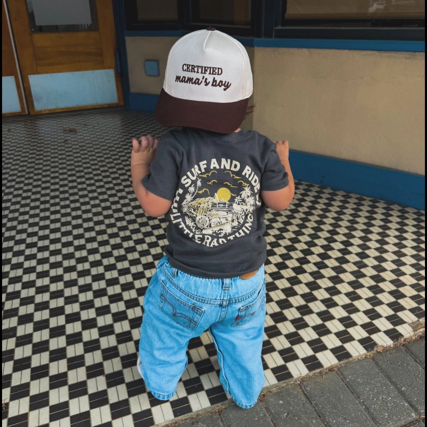 Child wearing a cap and t-shirt with text and graphics, standing on a checkered floor.