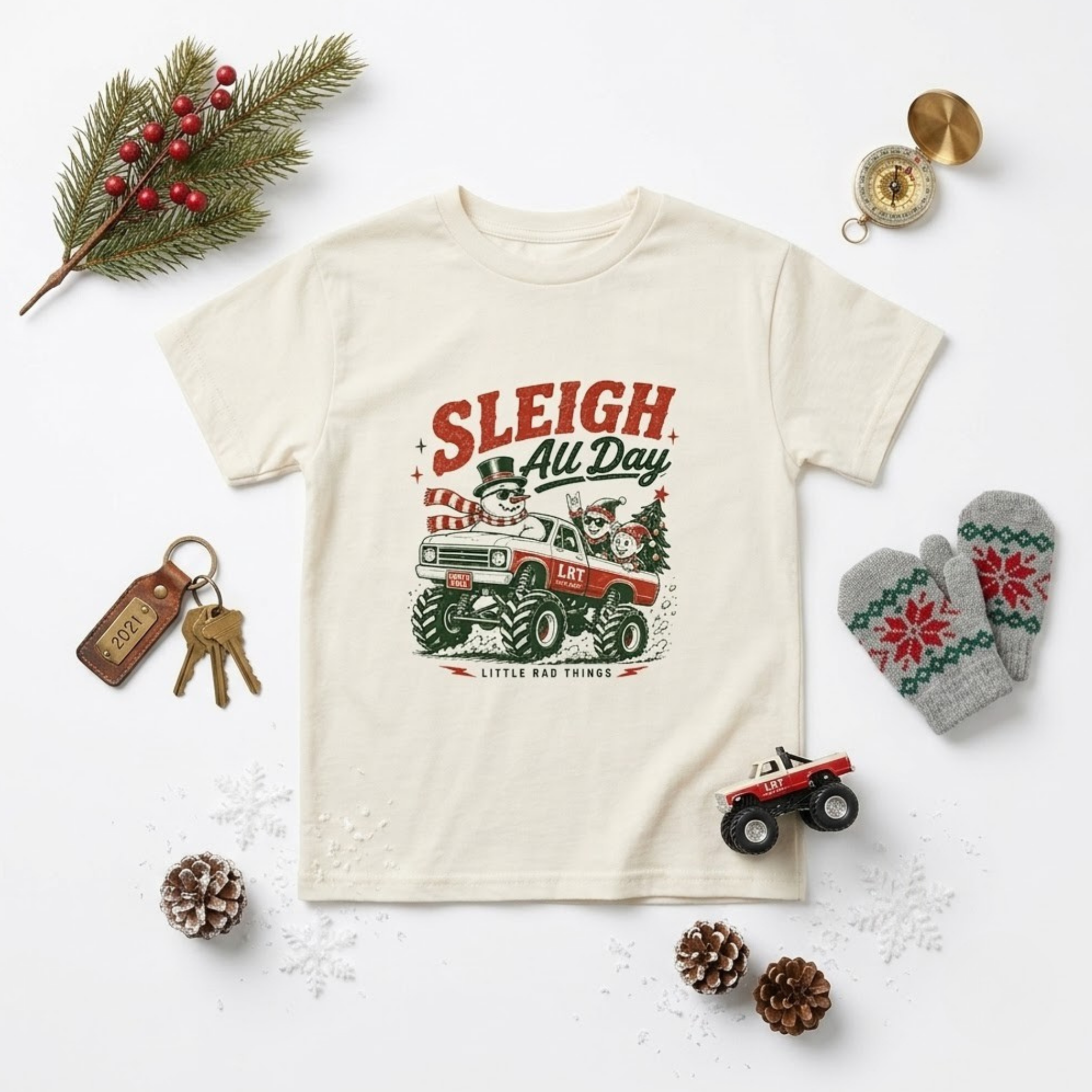 An off-white short-sleeve graphic t-shirt featuring a red and green tractor illustration with the text 'SLEIGH All Day' and 'Little Red Farm'. The t-shirt is styled in a flat-lay arrangement with Christmas-themed props including a pine branch with berries, a keychain, gray knit mittens, pinecones, and a small toy tractor on a white background.