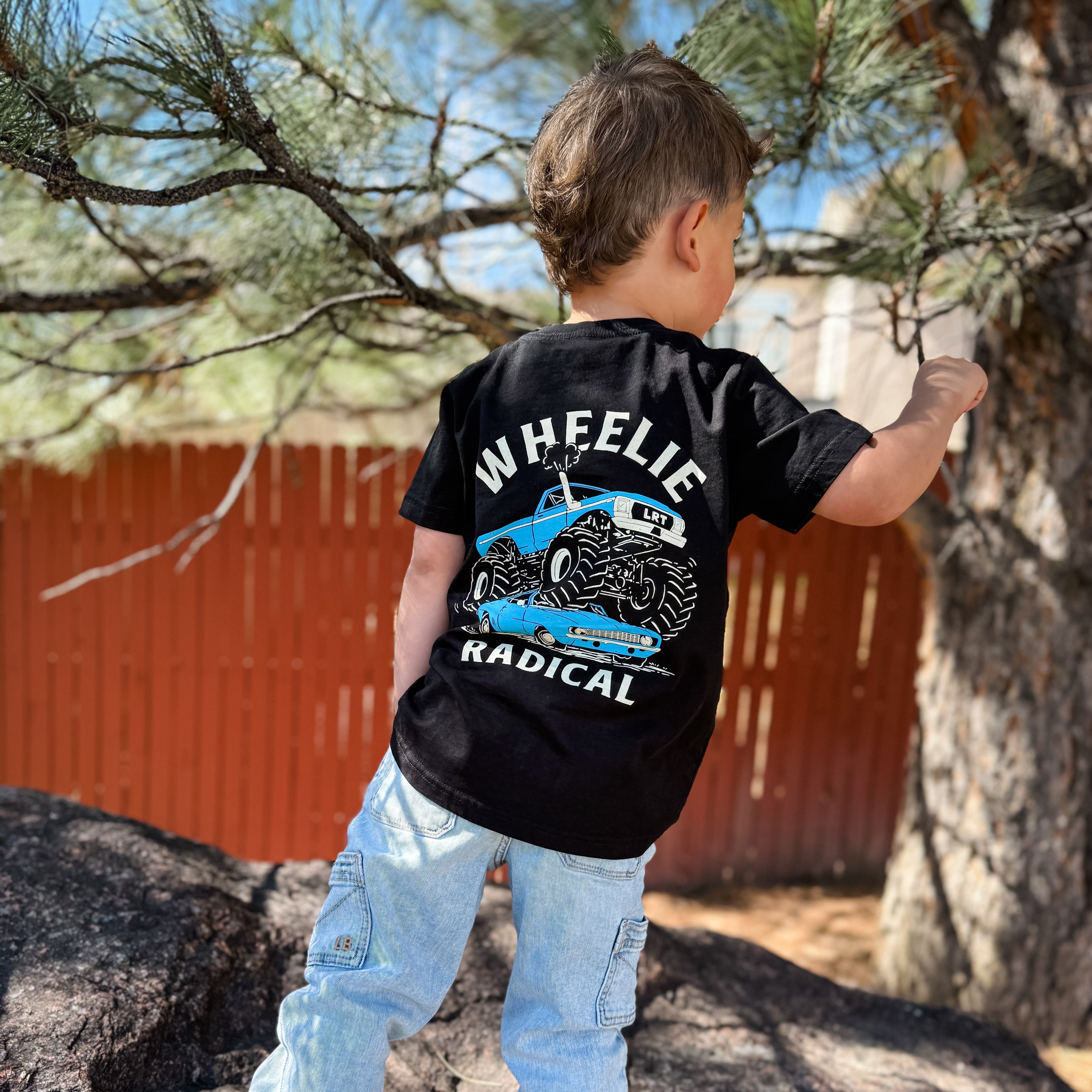 Child wearing a black t-shirt with graphic design and text, standing outdoors near trees and a red building.