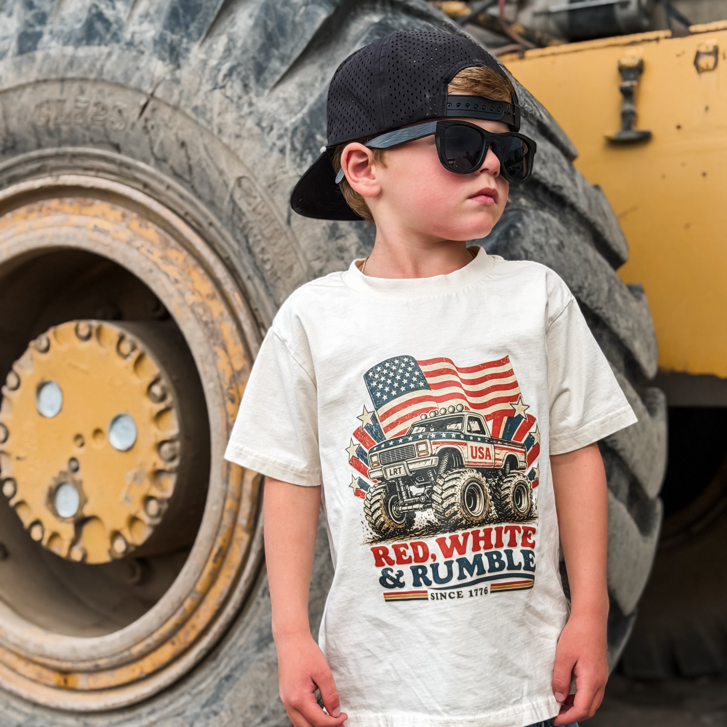 Child wearing a t-shirt with a graphic design of a truck and American flag, standing in front of a large construction vehicle.