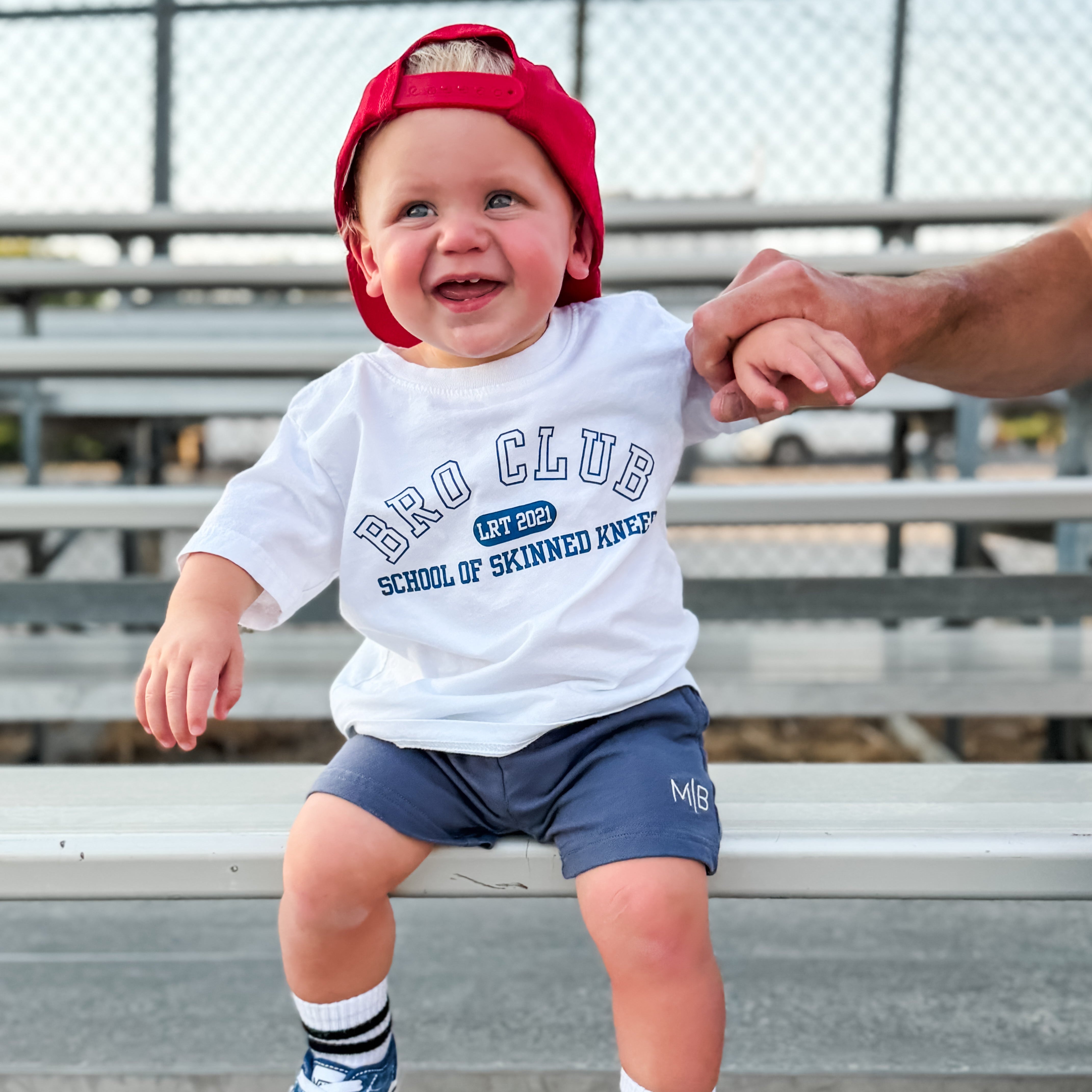 A toddler boy wearing a white 'Bro Club School of Skinned Knees' t-shirt, navy shorts, blue checkerboard sneakers, and a red cap sits on a metal bleachers at an outdoor sports field while holding an adult's hand.