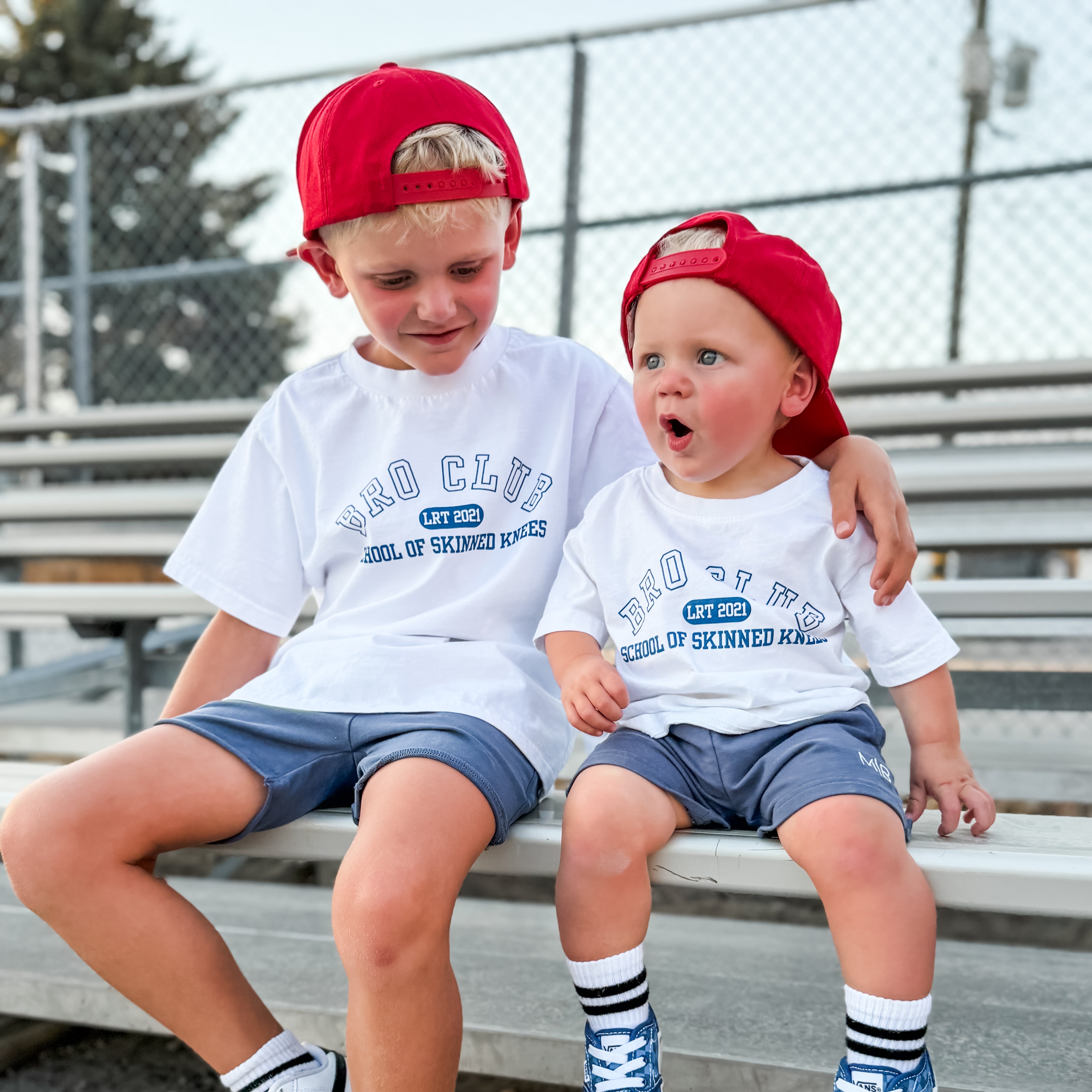 Two children sitting on metal bleachers outdoors, wearing matching white graphic T-shirts, blue shorts, red caps, and sneakers.