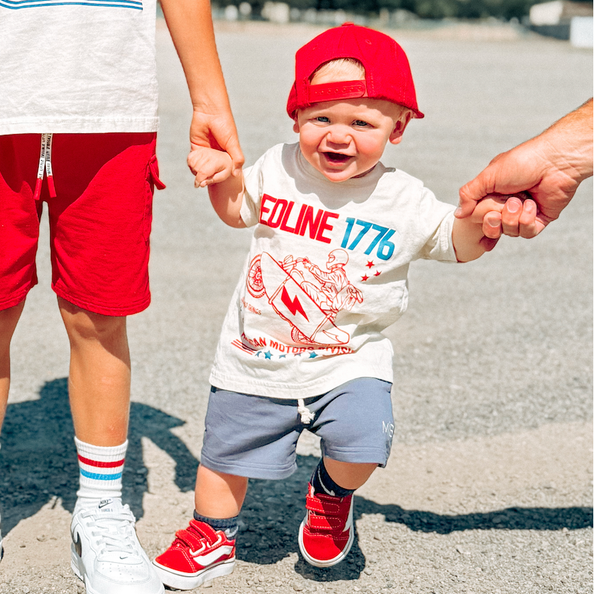 Two children wearing matching red and white outfits with sunglasses and hats, standing on a road with mountains in the background.