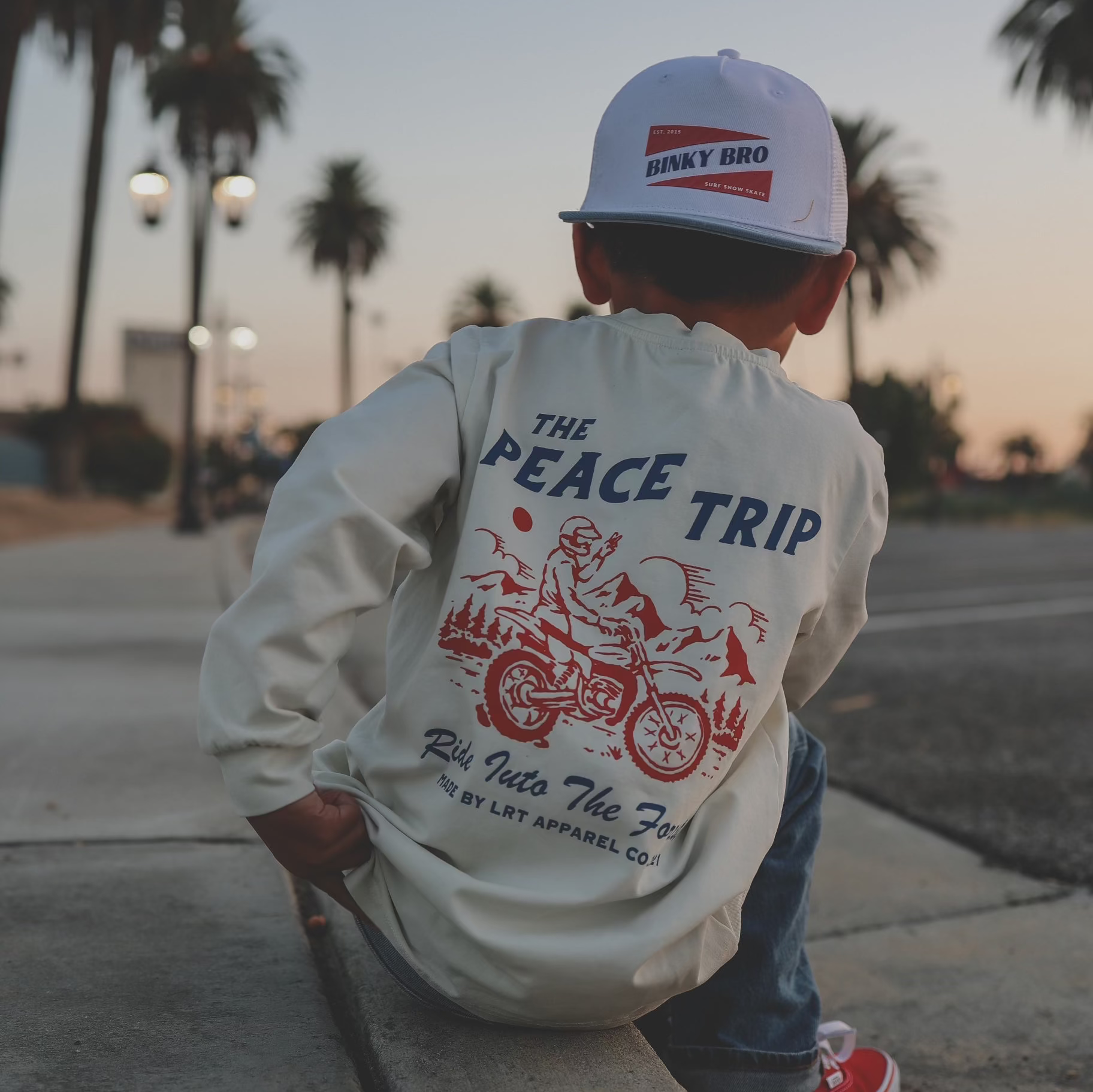 A child wearing a white long sleeve tee with red and blue checkered sleeves and a graphic print in the center, sitting on a sidewalk with palm trees in the background.