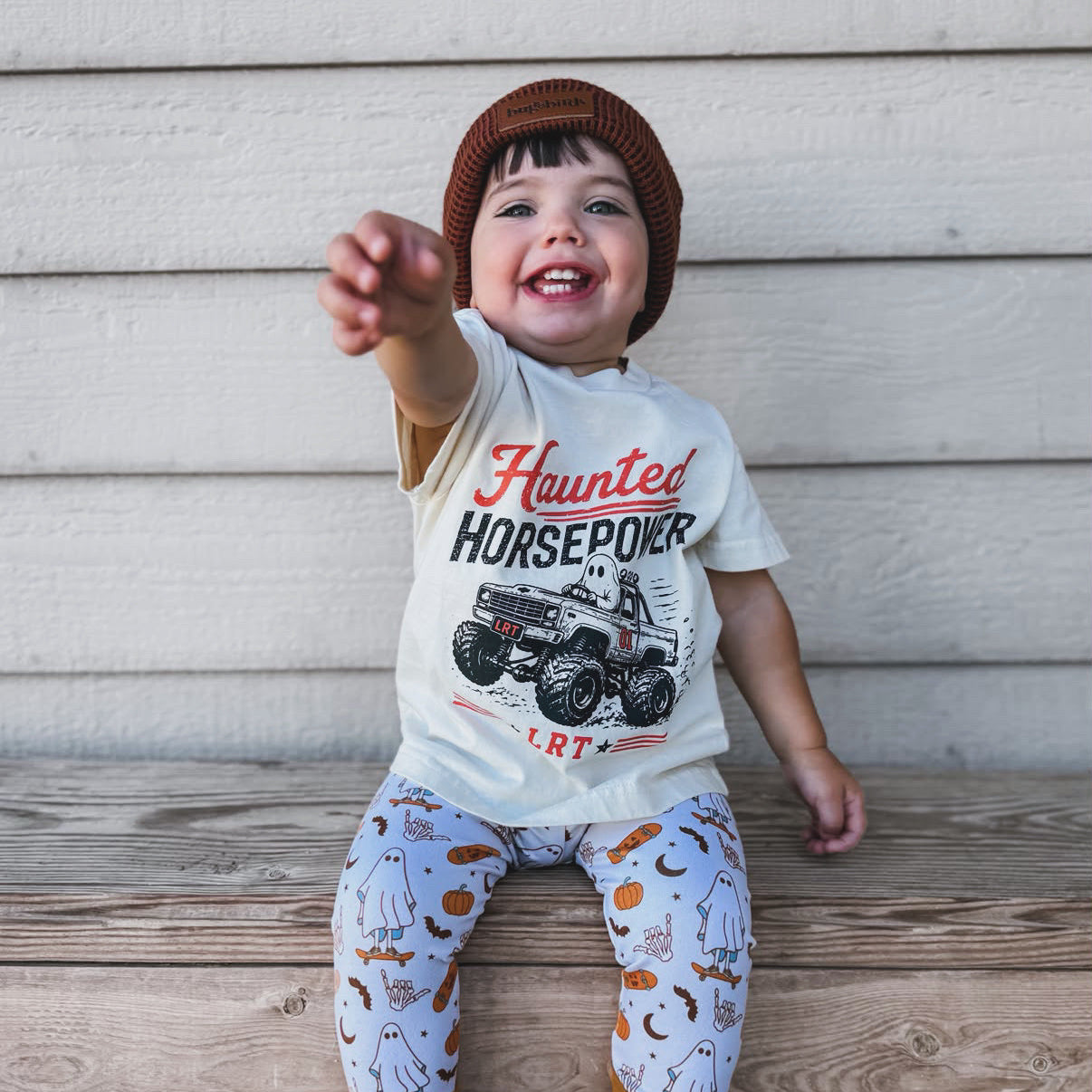 A smiling toddler sitting on a wooden bench against a light wood siding wall, wearing a cream graphic t-shirt that says 'Haunted Horsepower' with a monster truck, patterned leggings with brown leg cuffs, and a brown knit beanie.