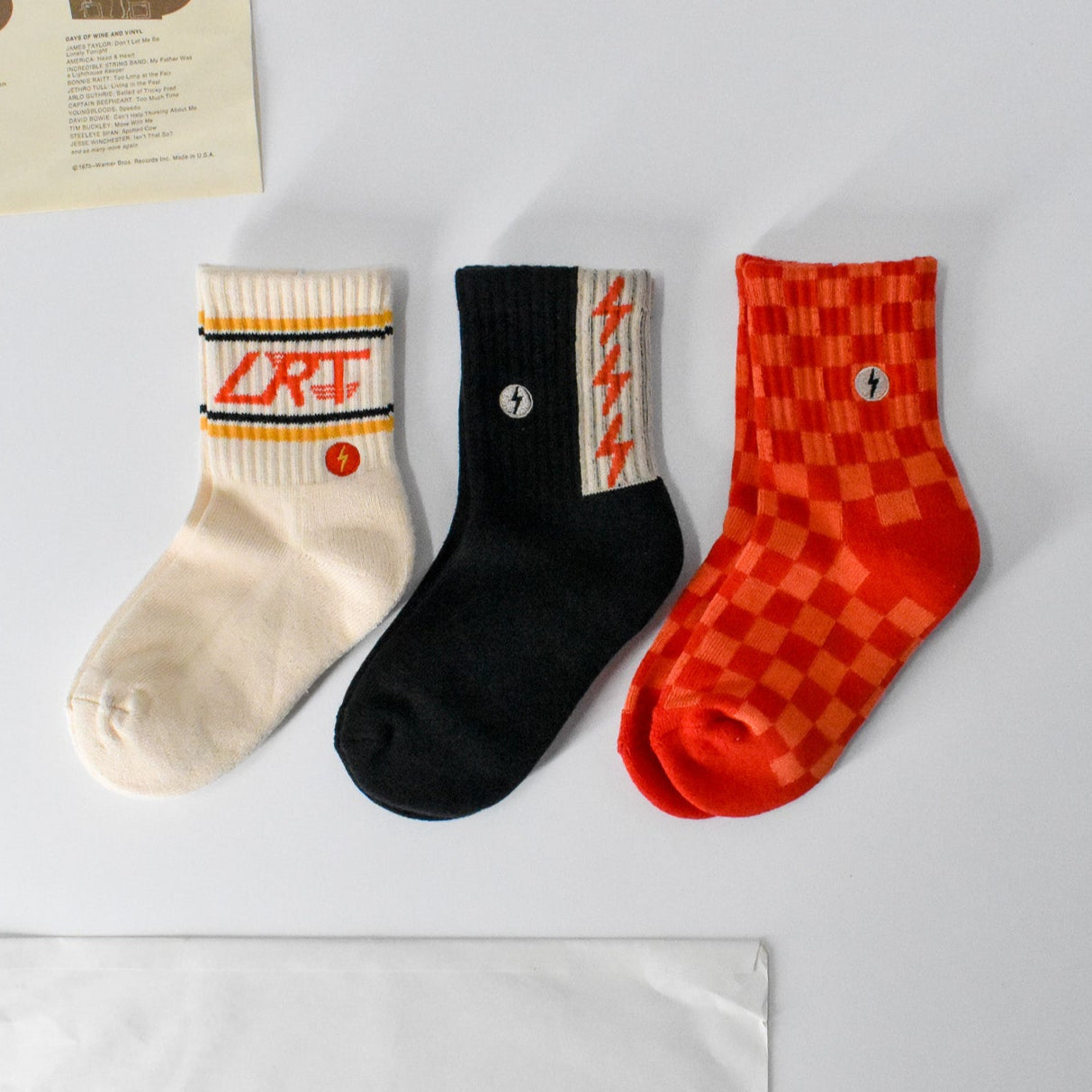 Three kids' socks displayed flat showing vintage-inspired designs - red/orange check pattern, funky spark print, and cream with LRT logo on ankle. Made from soft cotton blend."