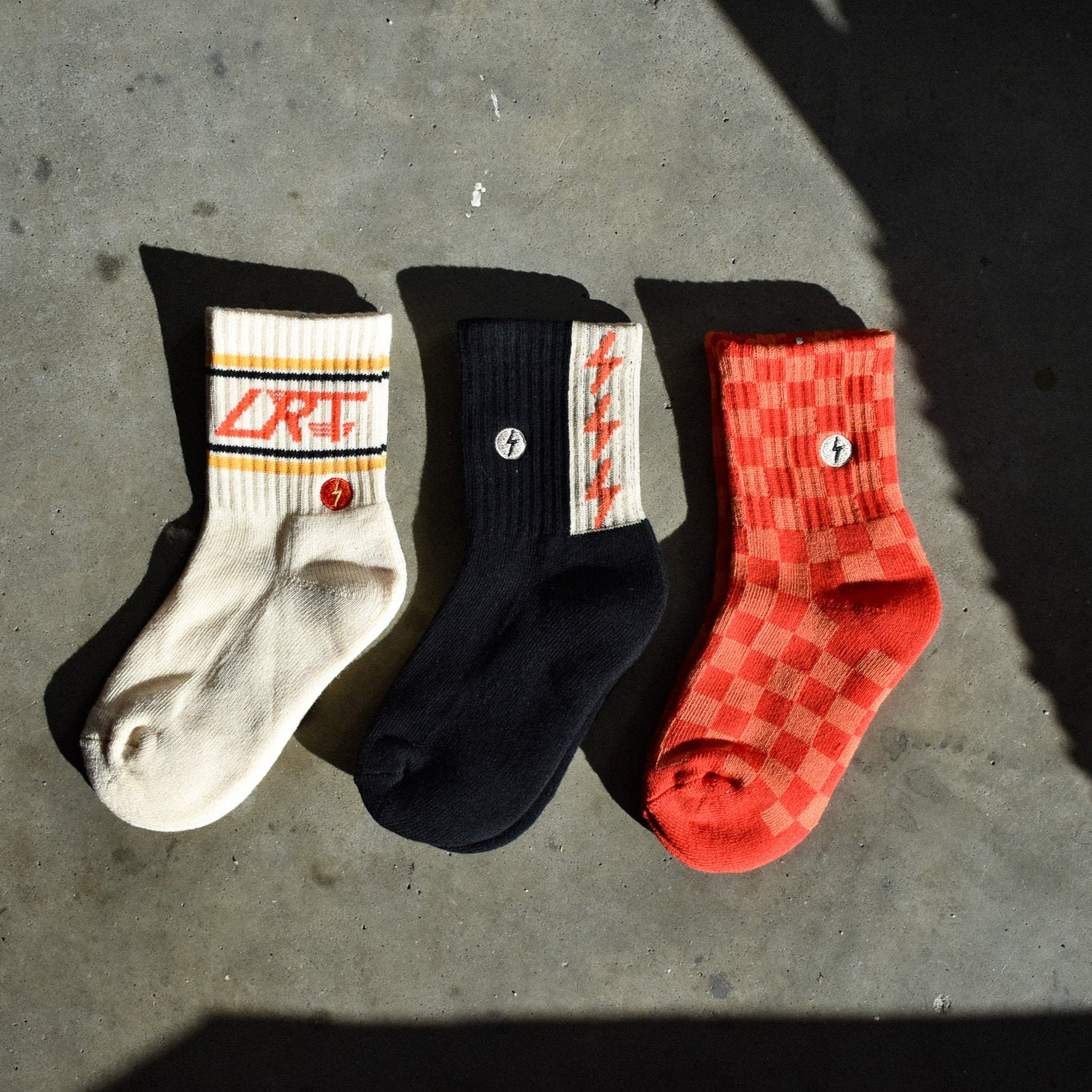 Three kids' socks displayed flat showing vintage-inspired designs - red/orange check pattern, funky spark print, and cream with LRT logo on ankle. Made from soft cotton blend."