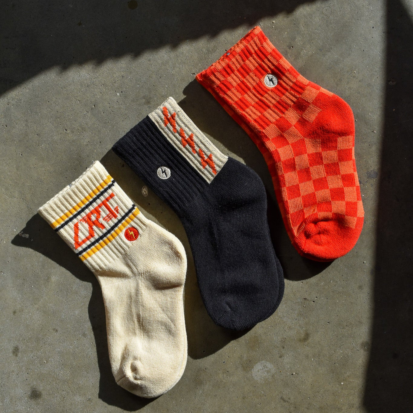 Three kids' socks displayed flat showing vintage-inspired designs - red/orange check pattern, funky spark print, and cream with LRT logo on ankle. Made from soft cotton blend."