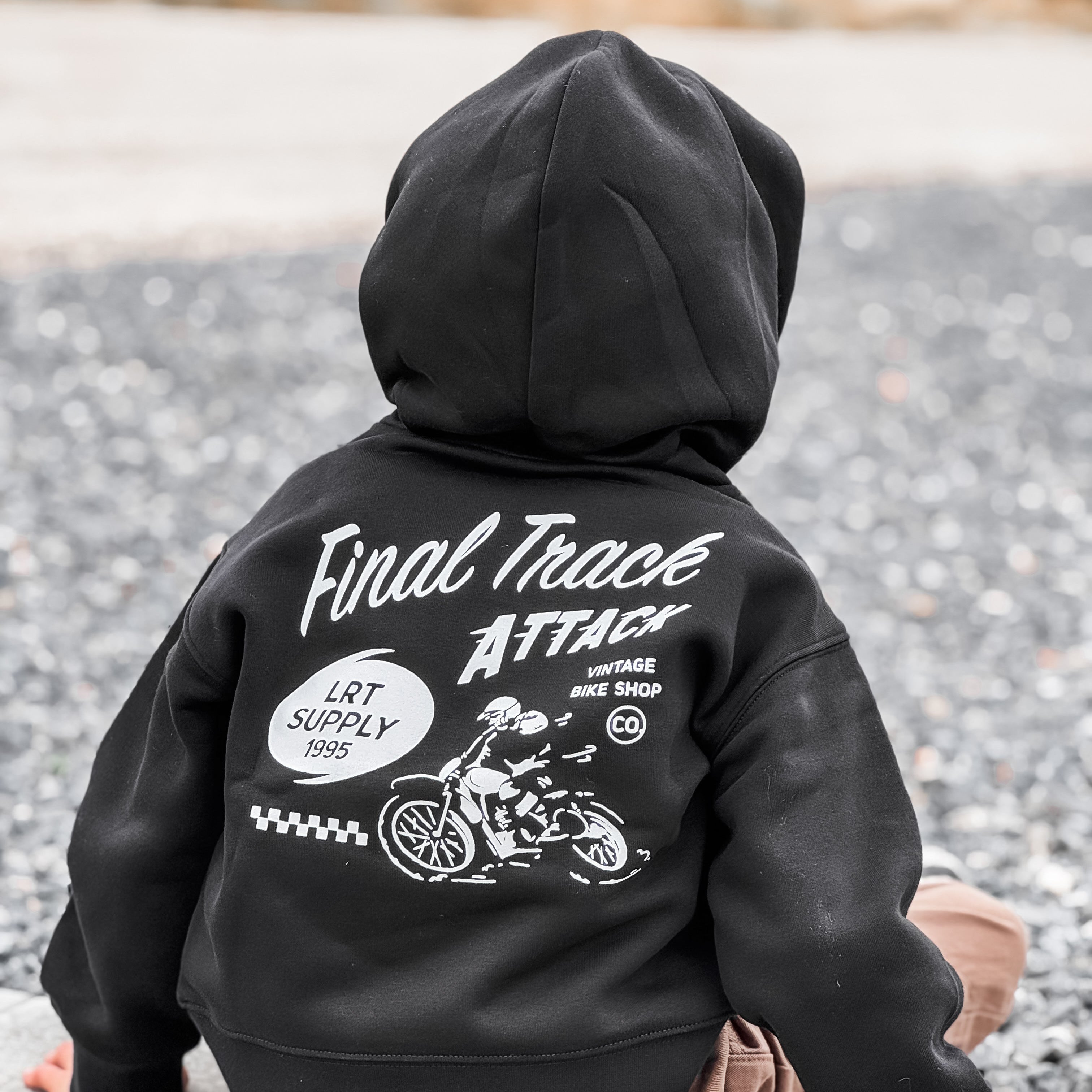 Child wearing a black hoodie with 'Final Trace Attack' design on the back, sitting on a pavement.