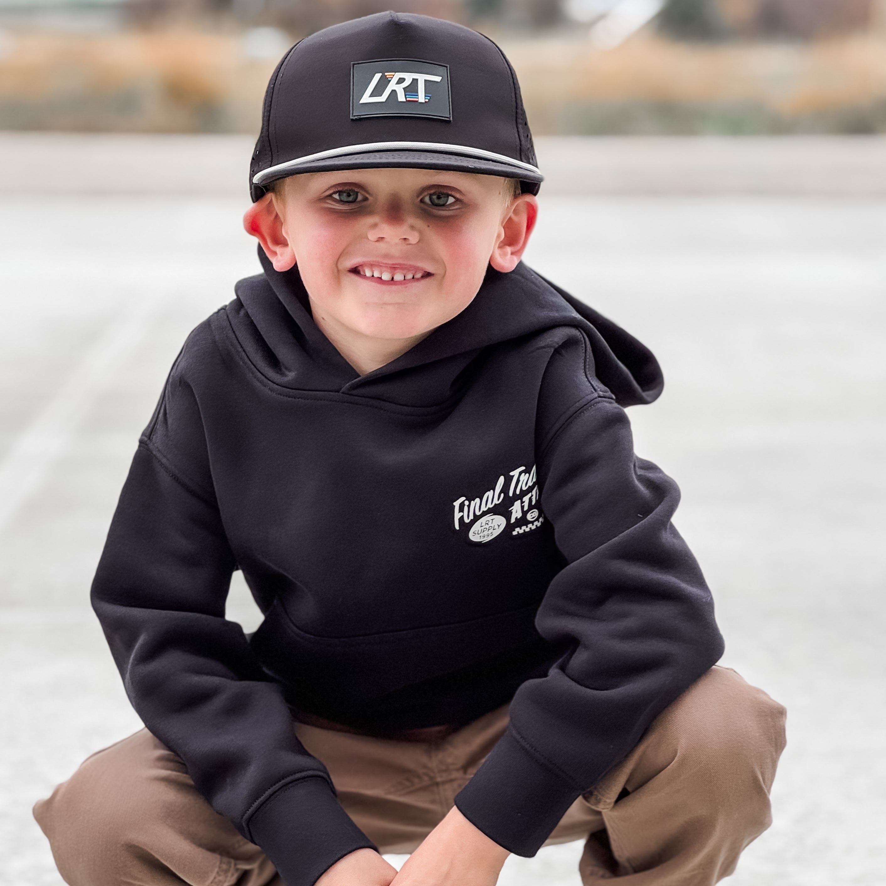 Child wearing a black hoodie and cap with visible branding, squatting on a road.
