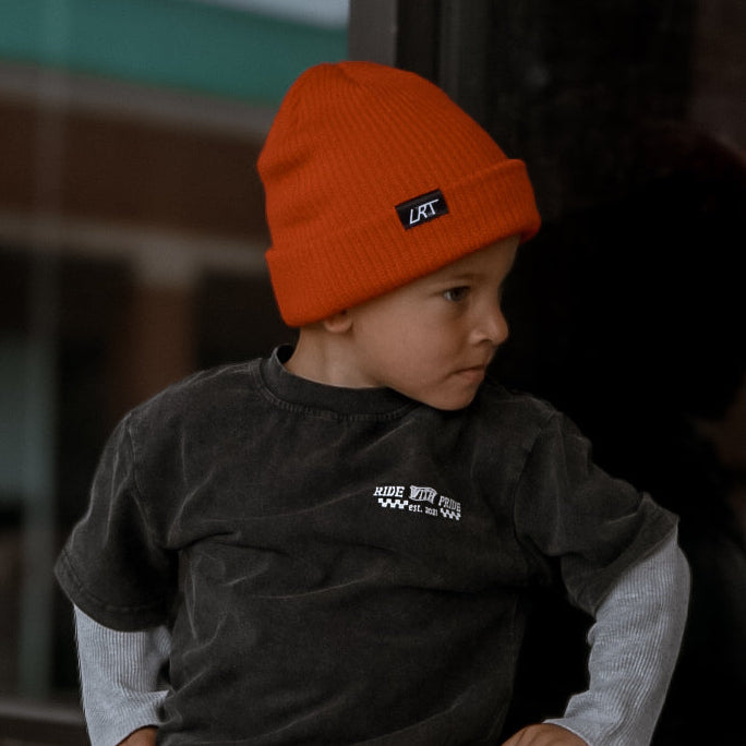 Child wearing an orange beanie and dark clothing leaning against a brick wall.