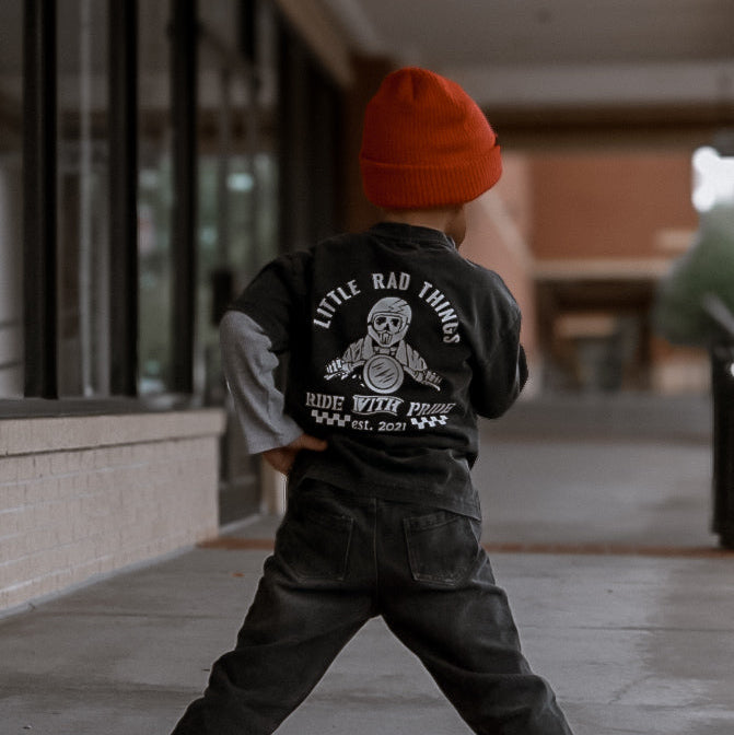Child wearing a black jacket with text and graphics, standing on a sidewalk.