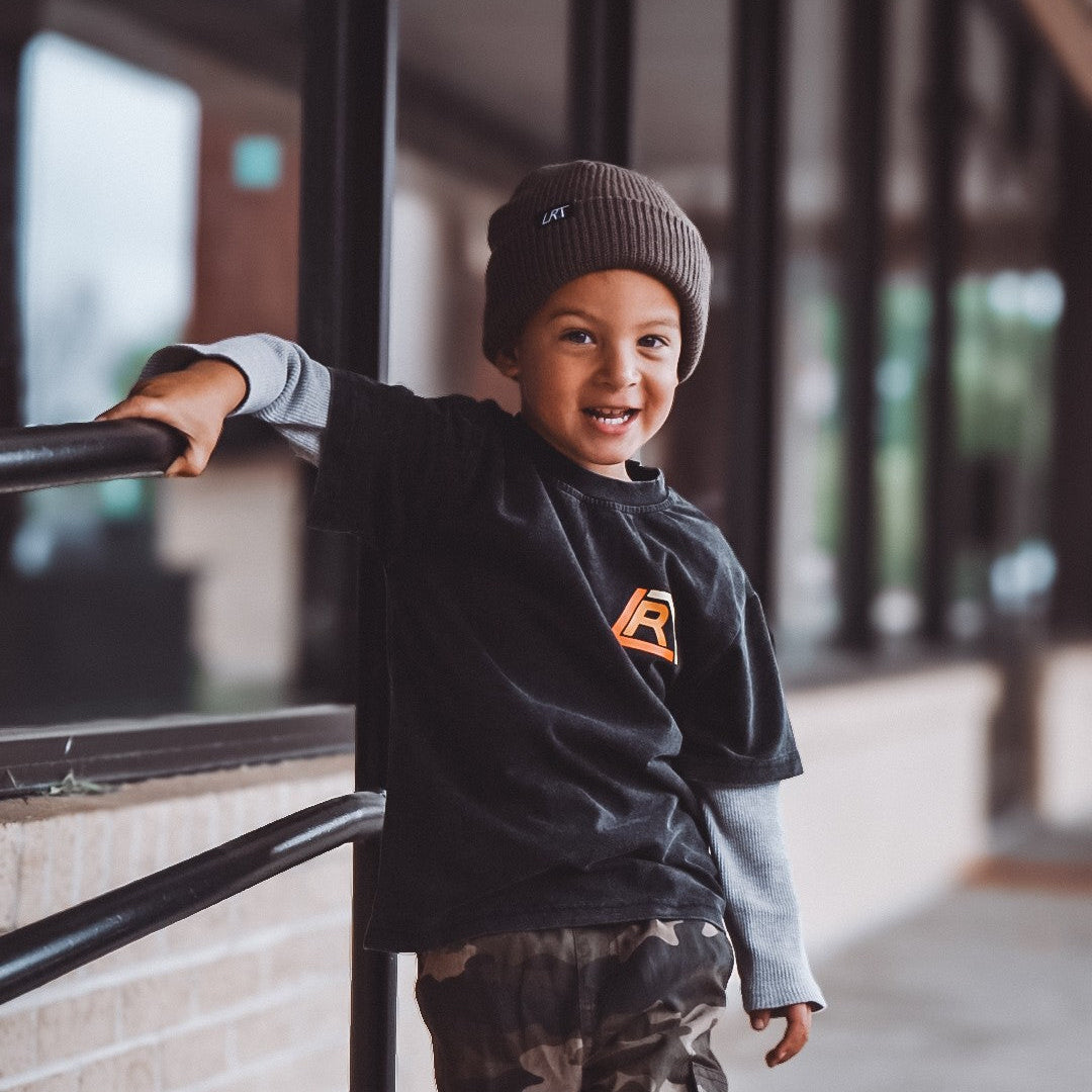 Child wearing a black sweatshirt with a logo, camouflage pants, and a beanie leaning against a railing indoors.